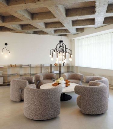 Um interior moderno com o candeeiro suspenso Veskt da Normann Copenhagen como elemento central que chama a atenção. O candeeiro escultural preto com vários braços curvos e lâmpadas suspensas ilumina uma mesa redonda rodeada por seis cadeiras estofadas. Tetos de betão bruto, linhas simples e cores suaves criam um ambiente harmonioso e contemporâneo.
