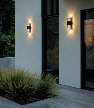 Parede exterior moderna com duas luzes de parede Aludra Double da Nordlux. As luzes iluminam para cima e para baixo, criando um jogo de luz elegante e acolhedor na fachada clara. Em frente à casa, gramíneas ornamentais verdes e linhas simples criam um ambiente arquitetónico elegante.