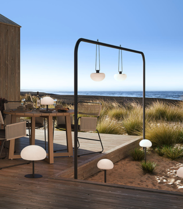 Candeeiros de exterior num terraço de madeira junto ao mar, luz suave entre a relva das dunas.