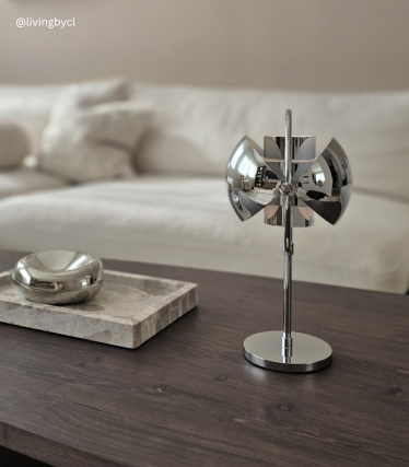 Candeeiro de mesa de design cromado sobre uma mesa de madeira em frente a um sofá de cor clara.