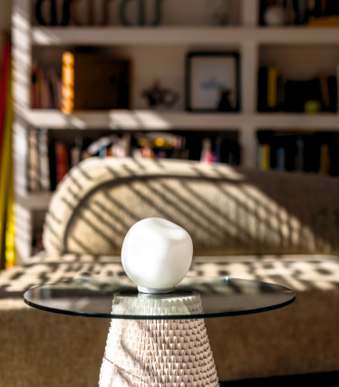 Pequeno candeeiro de mesa branco com tampo de vidro, luz solar e estante ao fundo.