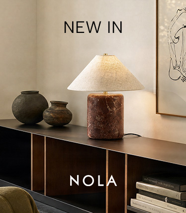 Candeeiro de mesa Nola com base em mármore castanho-avermelhado e abajur cónico em linho sobre um aparador escuro. Texto: «Novidade – Nola».