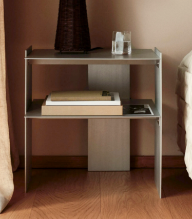 Mesa de cabeceira estreita em metal com duas prateleiras, livros, um copo e uma jarra junto à cama.