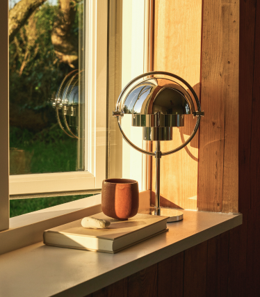 Candeeiro de mesa cromado no parapeito de uma janela à luz do entardecer, junto a um livro e uma caneca de cerâmica.