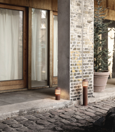 Cena exterior com duas luzes cilíndricas modernas colocadas ao lado de um caminho de pedra e uma coluna de tijolo e betão. Atrás delas, há portas de vidro com caixilhos de madeira e cortinas de luz, e um grande vaso com uma planta perene. O brilho acolhedor das luzes cria uma atmosfera aconchegante e convidativa.
