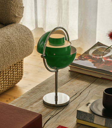 Candeeiro de mesa Multi-Lite verde com base cromada sobre uma mesa de madeira junto a livros.