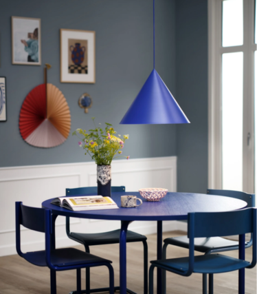 Candeeiro suspenso azul em forma de cone sobre uma mesa de jantar redonda azul com um ramo de flores, numa sala de jantar com parede azul e obras de arte.